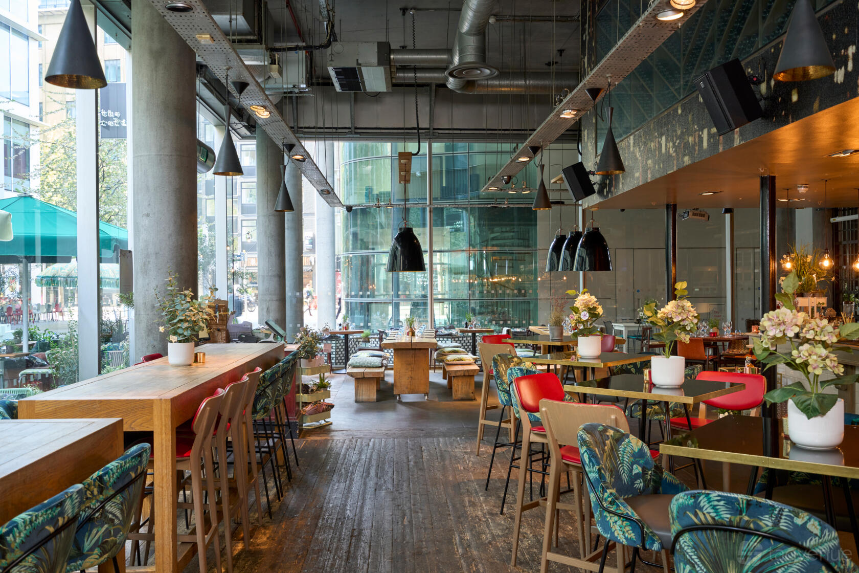 A restaurant with floor-to-ceiling windows, wooden tables, colorful chairs, and pendant lighting at The Refinery Bankside.