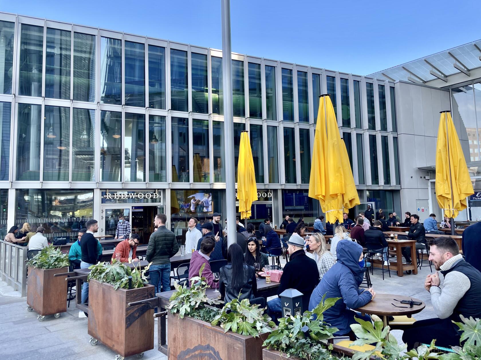 A pub terrace with wooden tables, yellow umbrellas, and planters in front of large glass windows at Redwood.