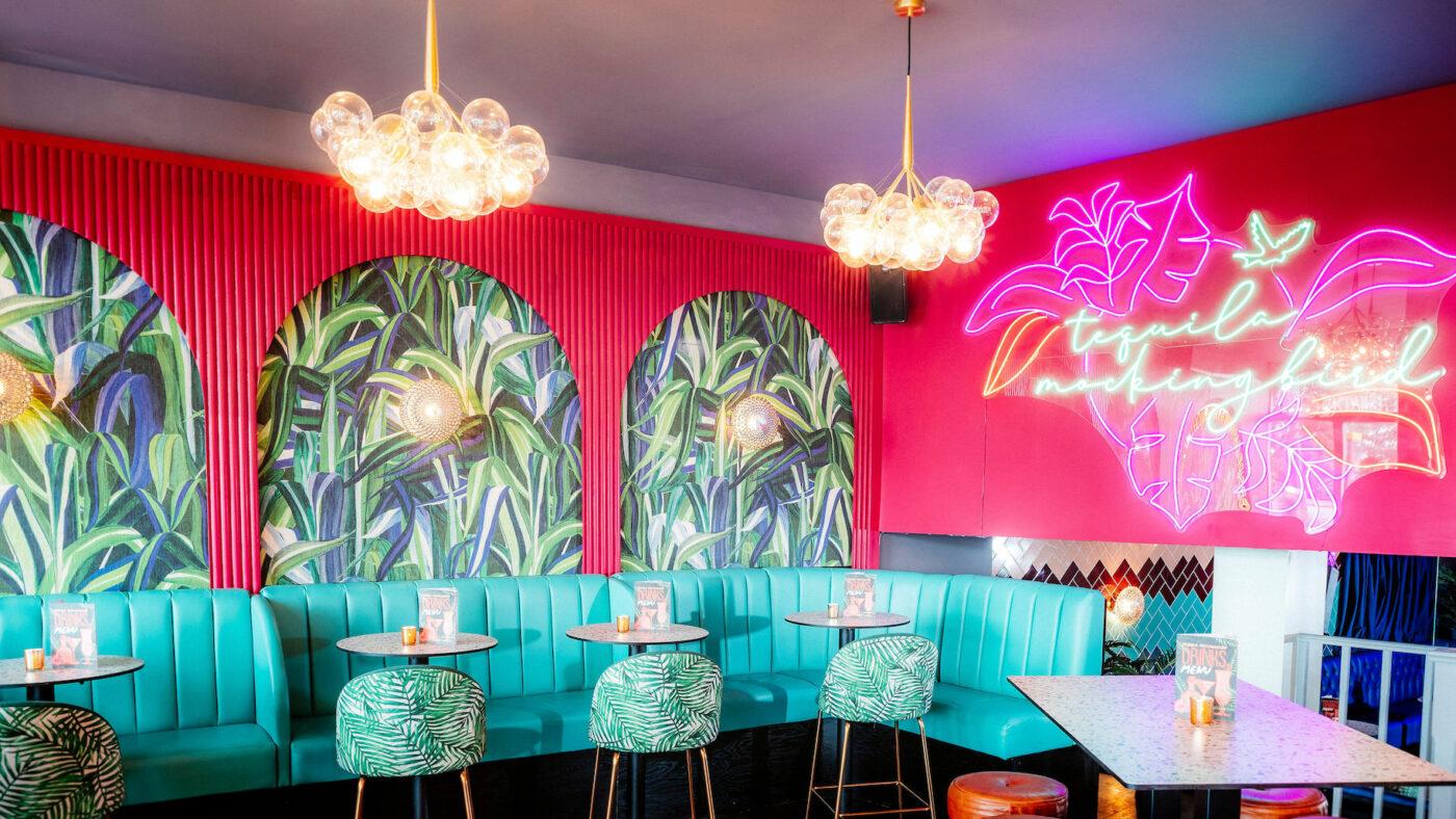A cocktail bar with teal banquette seating, tropical wallpaper, and neon wall art at Tequila Mockingbird Putney.
