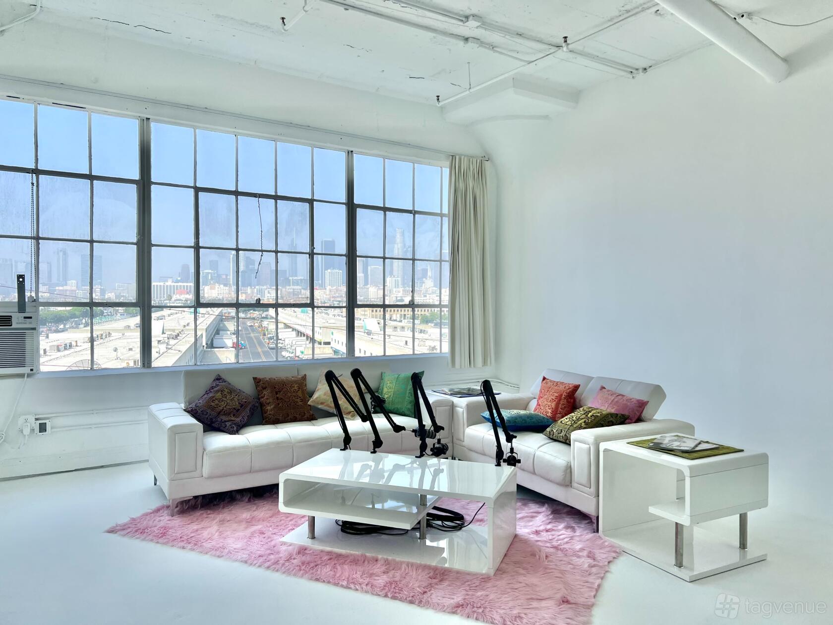 A podcast studio with large industrial windows, white furniture, colorful pillows, and microphones at Alameda Tower.