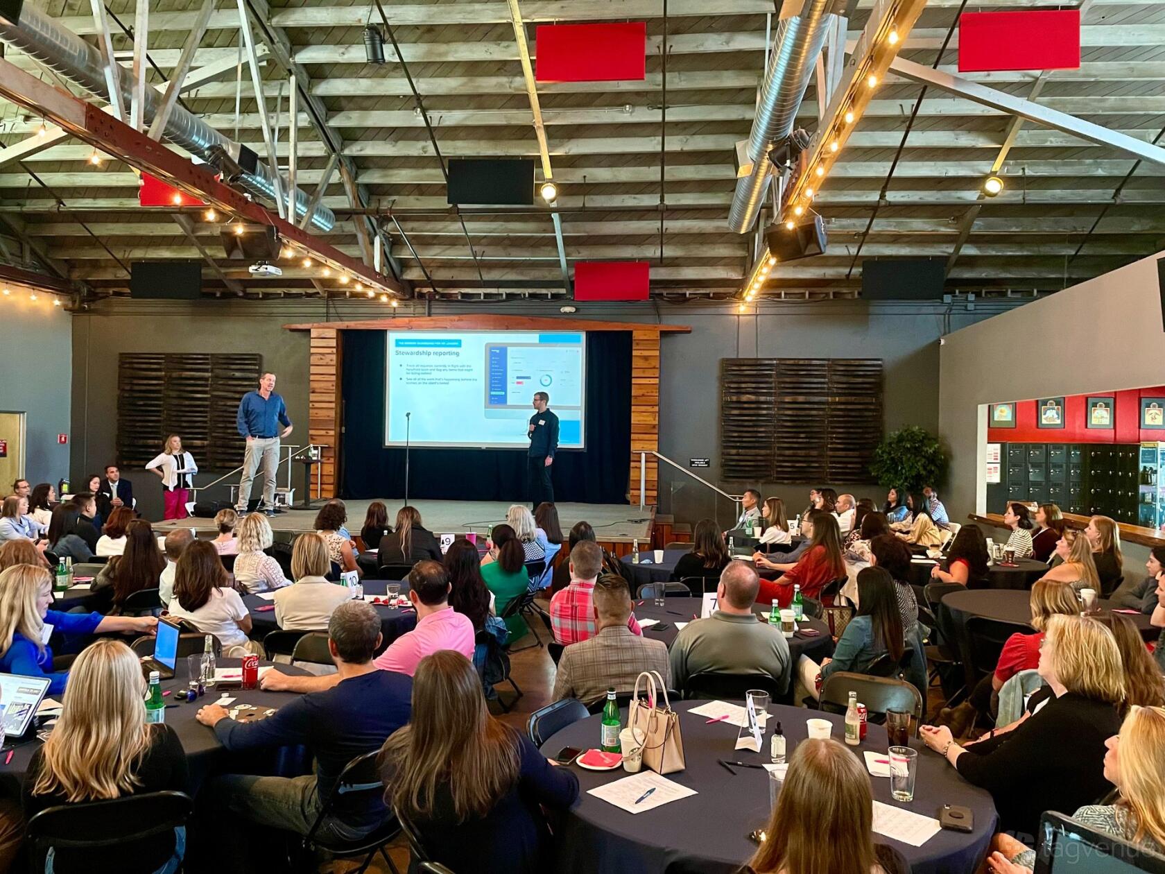 A warehouse hall with exposed beams, ductwork, round tables, and a stage with a projector screen at Devil's Canyon Brewing Company.