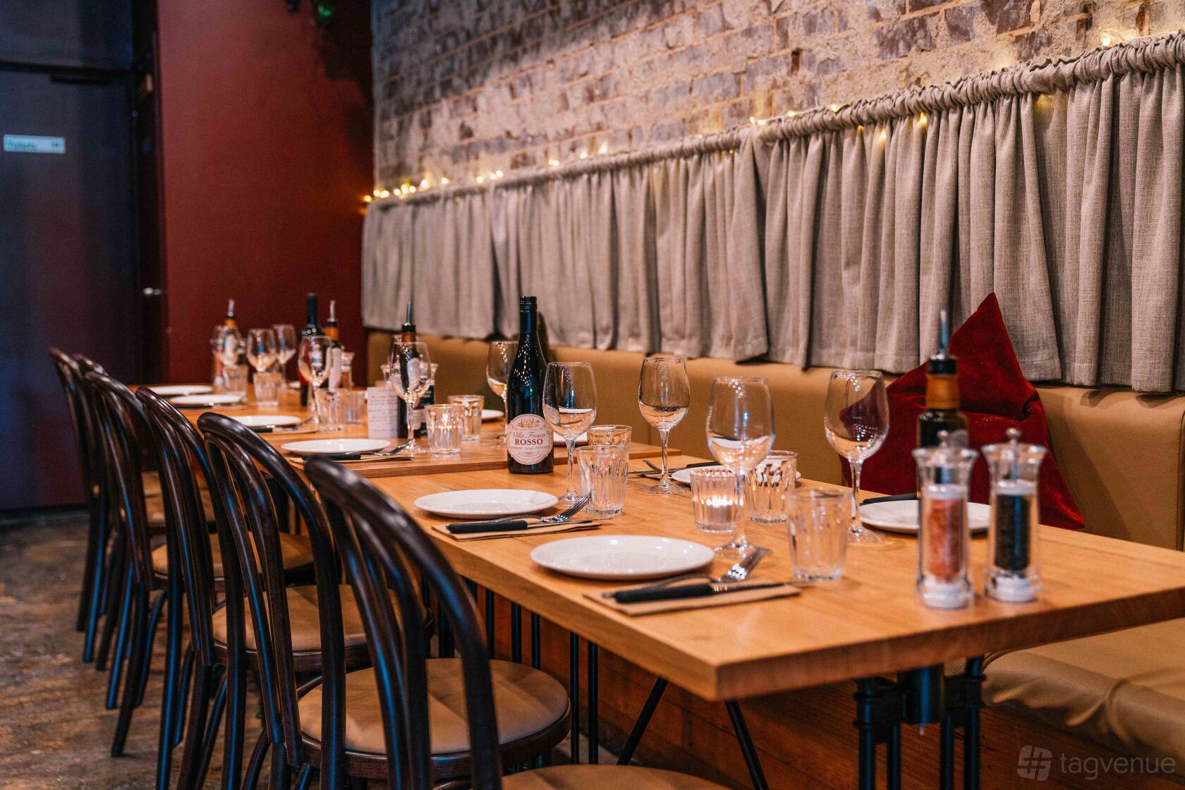 A restaurant dining area with a long wooden table, brick walls, wine bottles, and cushioned bench seating at Farro Hawthorn.
