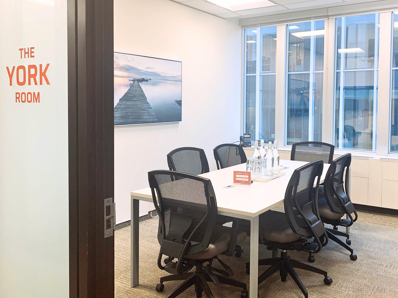 A meeting room with glass windows, a long table, mesh chairs, and bottled water at The Professional Centre.