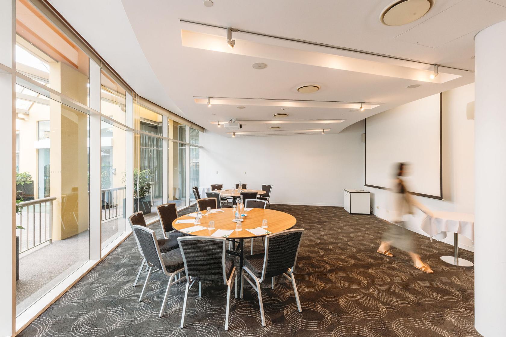 A meeting room with round tables, black chairs, carpeted floors, and floor-to-ceiling windows at Holiday Inn Sydney Potts Point.