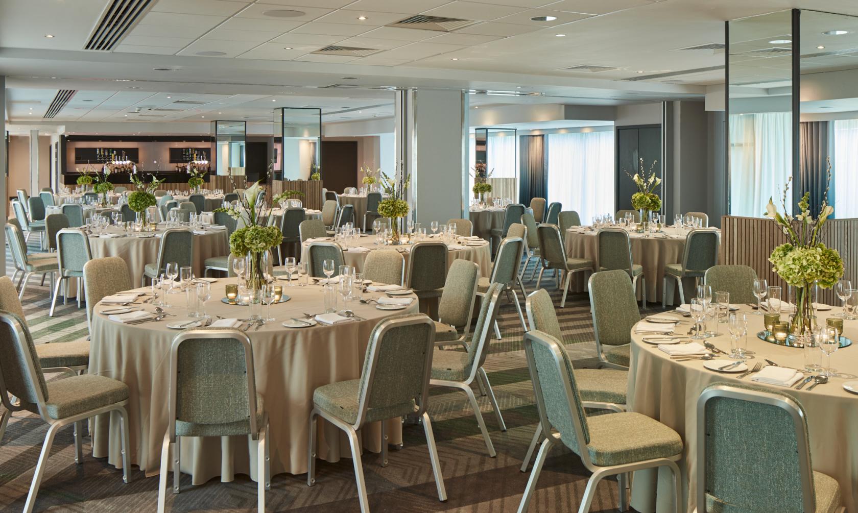A banquet hall with round tables set with white linens, glassware, and green floral centerpieces at Manchester Marriott Hotel Piccadilly.