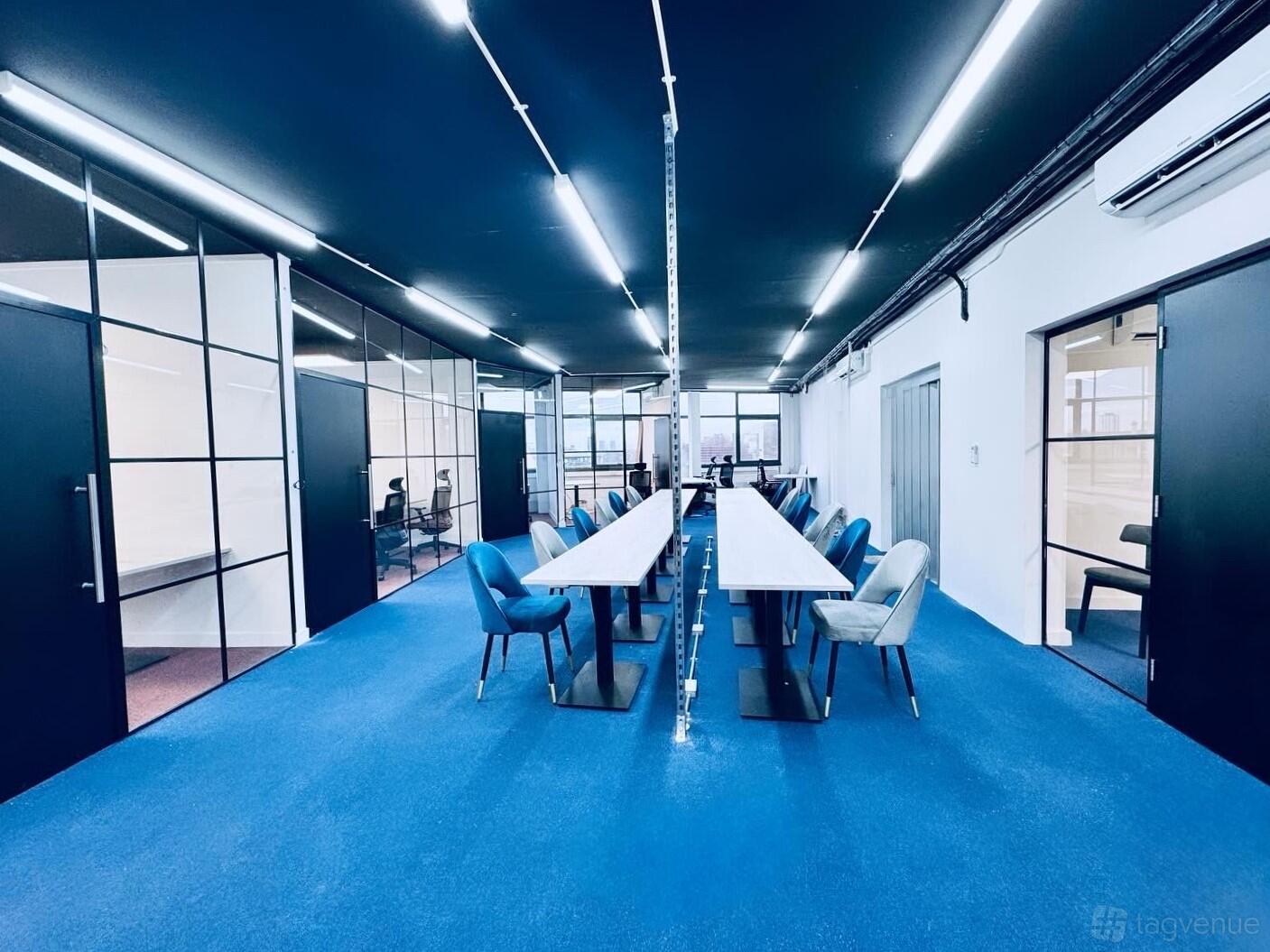 A coworking meeting room with blue carpet, glass partition walls, and long white desks at The Factory London.