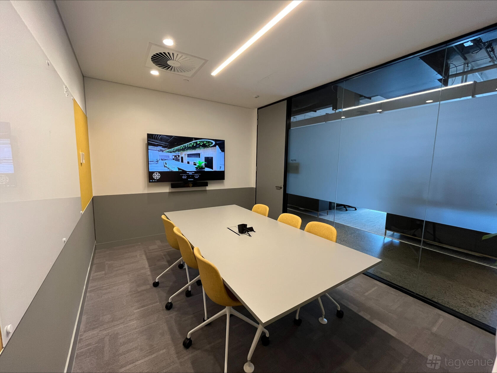 A coworking space meeting room with glass walls, a wall-mounted screen, and yellow chairs at cirque South Brisbane.