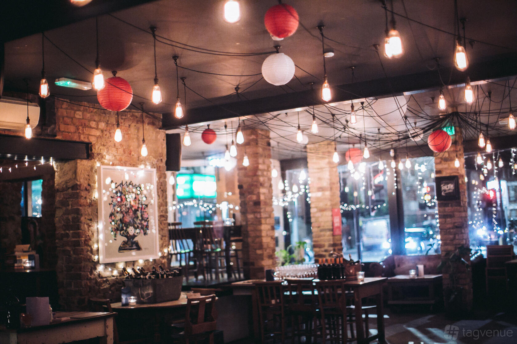 A bar with exposed brick walls, hanging Edison bulbs, and red lanterns at The Alice House Queen's Park.