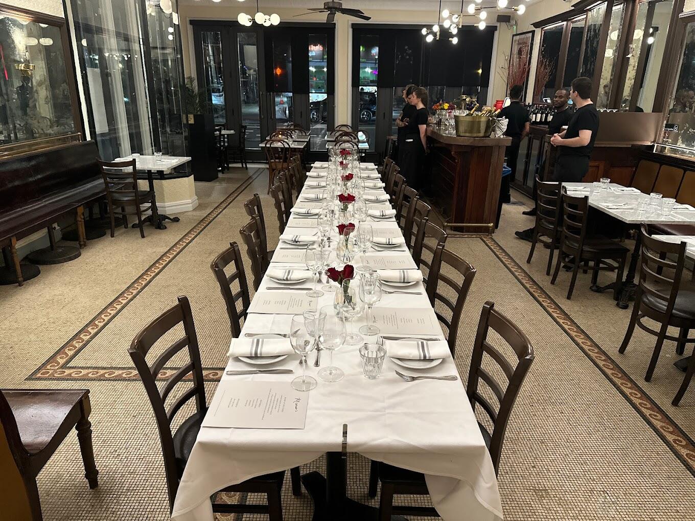 A restaurant with long banquet tables set with white linens, menus, and red rose centerpieces at Brasserie Le Mistral.