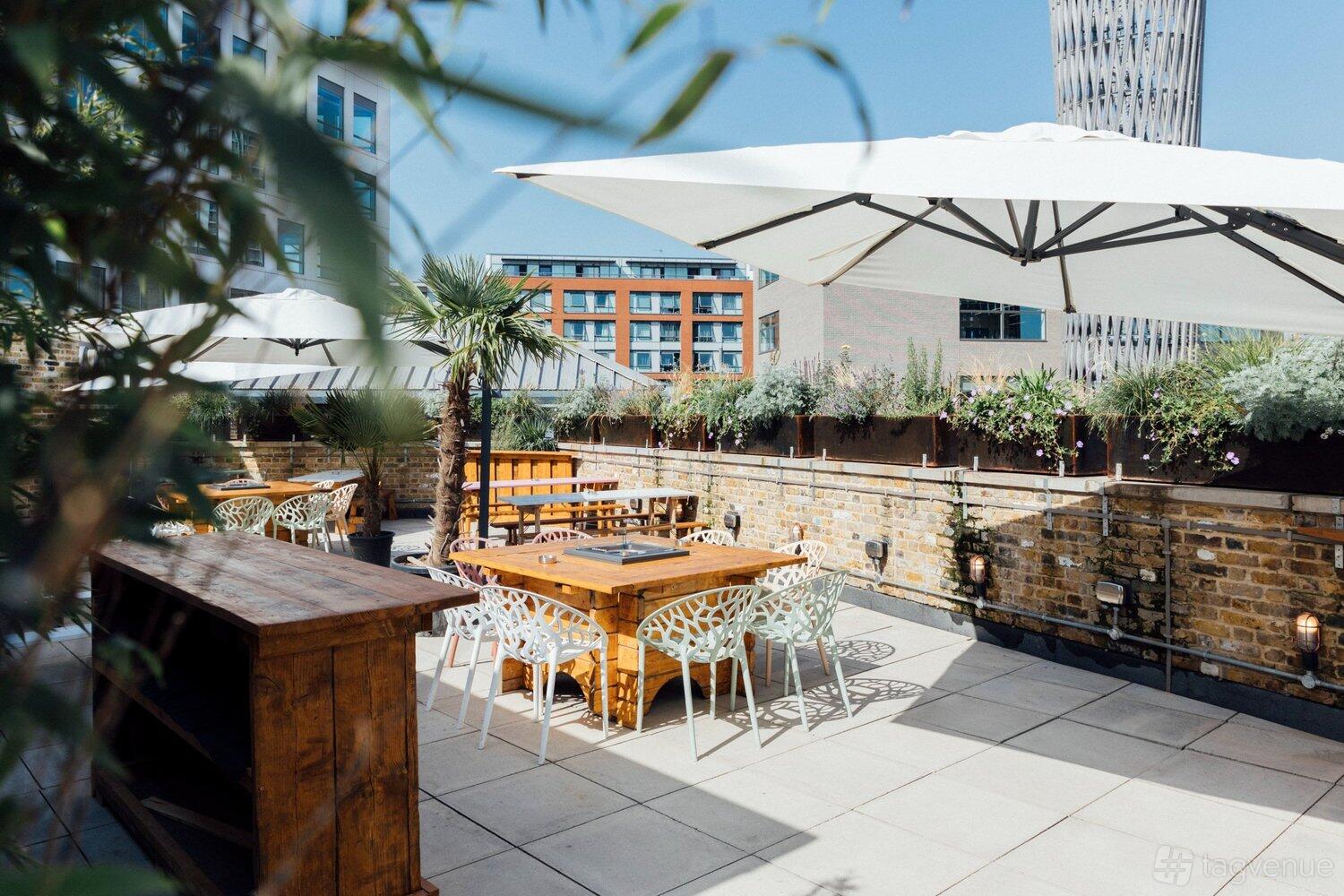 A rooftop bar with wooden tables, white chairs, large umbrellas, and brick walls at The Rum House at Jim & Tonic East.