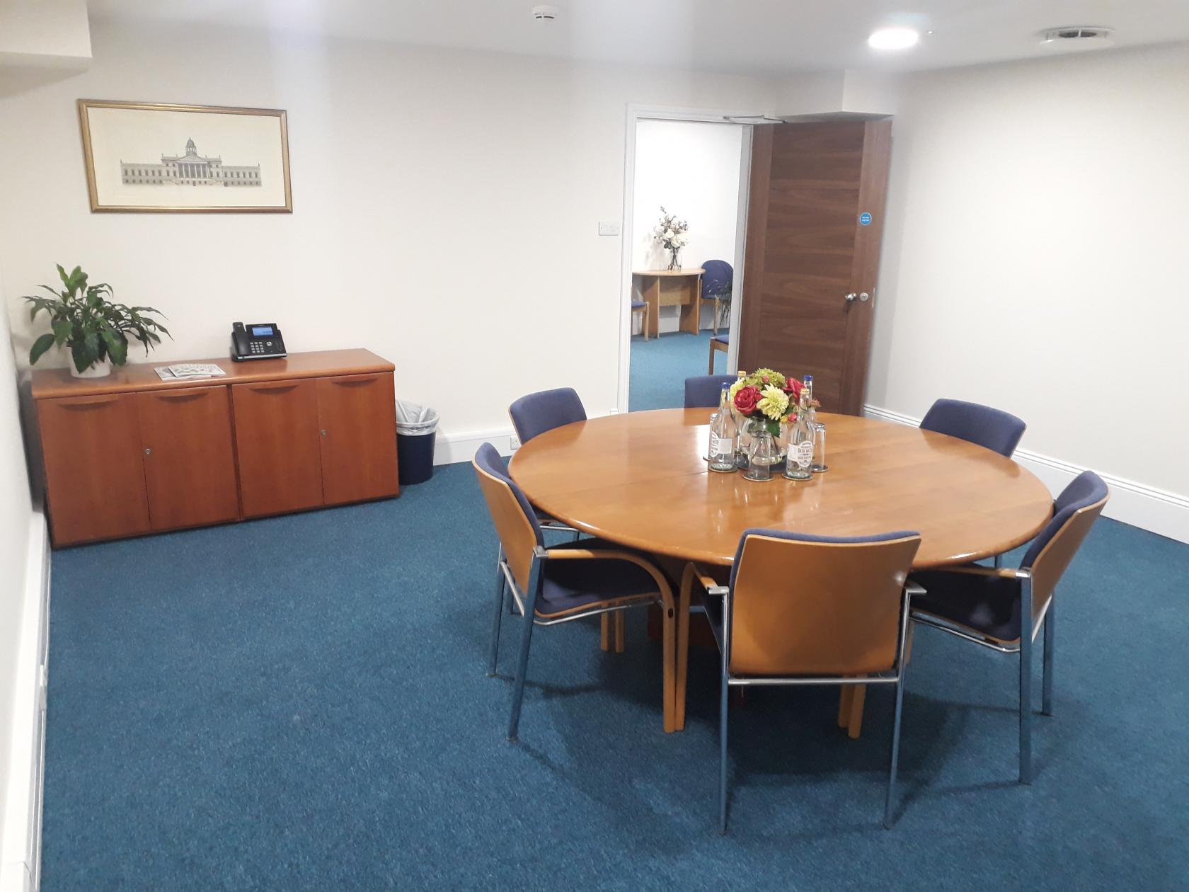 A meeting room with a round wooden table, six chairs, blue carpet, and a sideboard at Inigo Business Centres Bath.