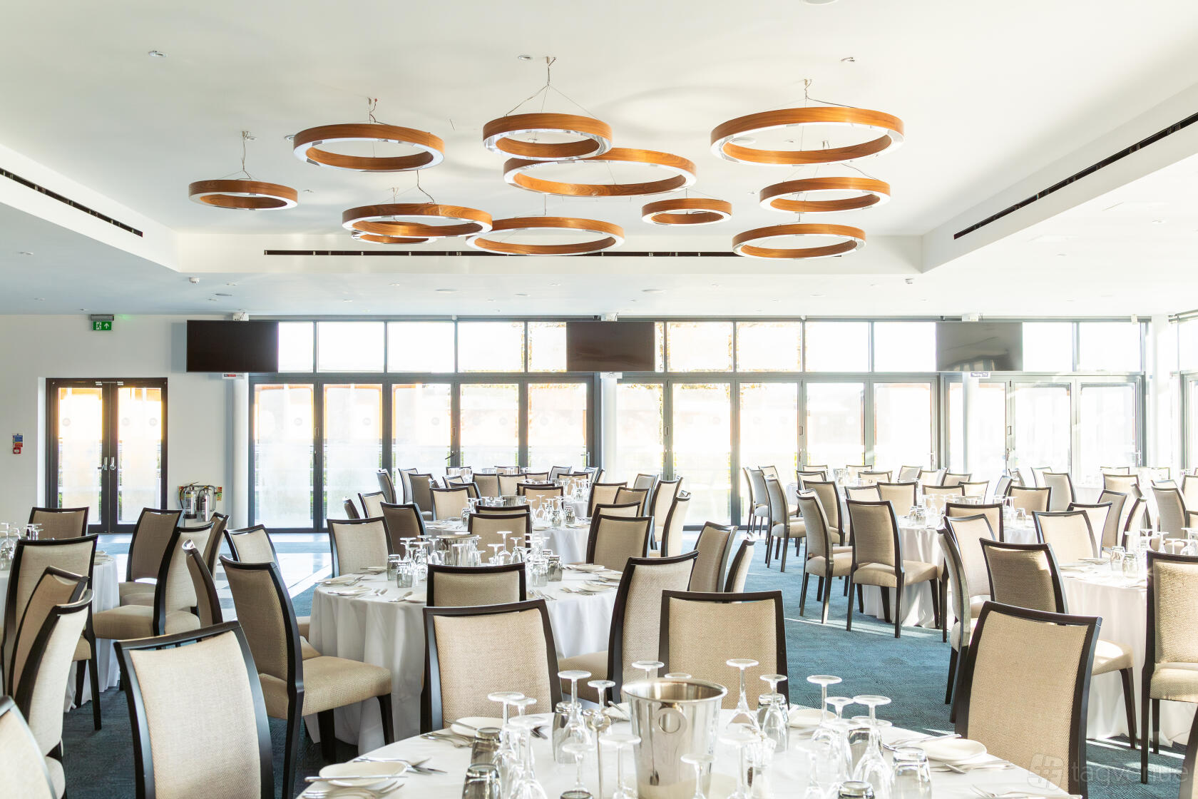 An event venue with round tables, beige upholstered chairs, large windows, and modern circular light fixtures at Newbury Racecourse.