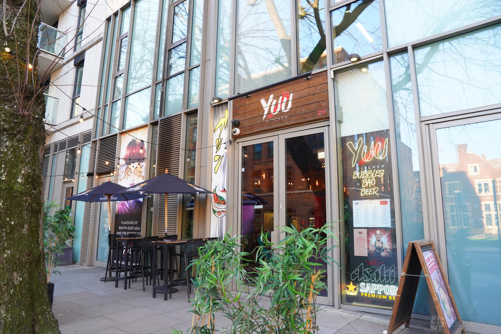 A restaurant with floor-to-ceiling windows, outdoor seating under black umbrellas, and menu boards at YUU Spitalfields
