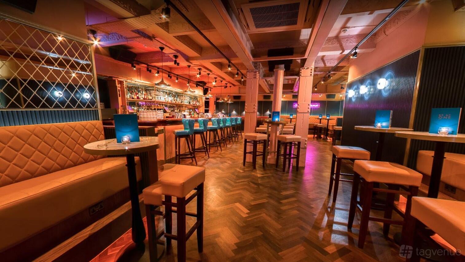 A cocktail bar with a long polished counter, leather banquette seating, high tables, and neon lighting at Be At One Birmingham - New Street.