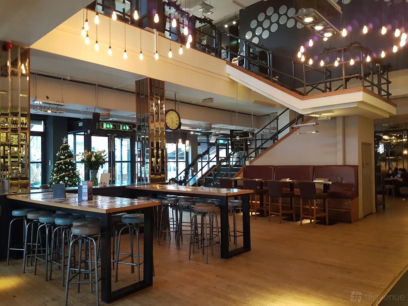 An event space in a bar with hanging pendant lights, wooden high tables, and a staircase leading to a mezzanine at All Bar One Brindley Place.