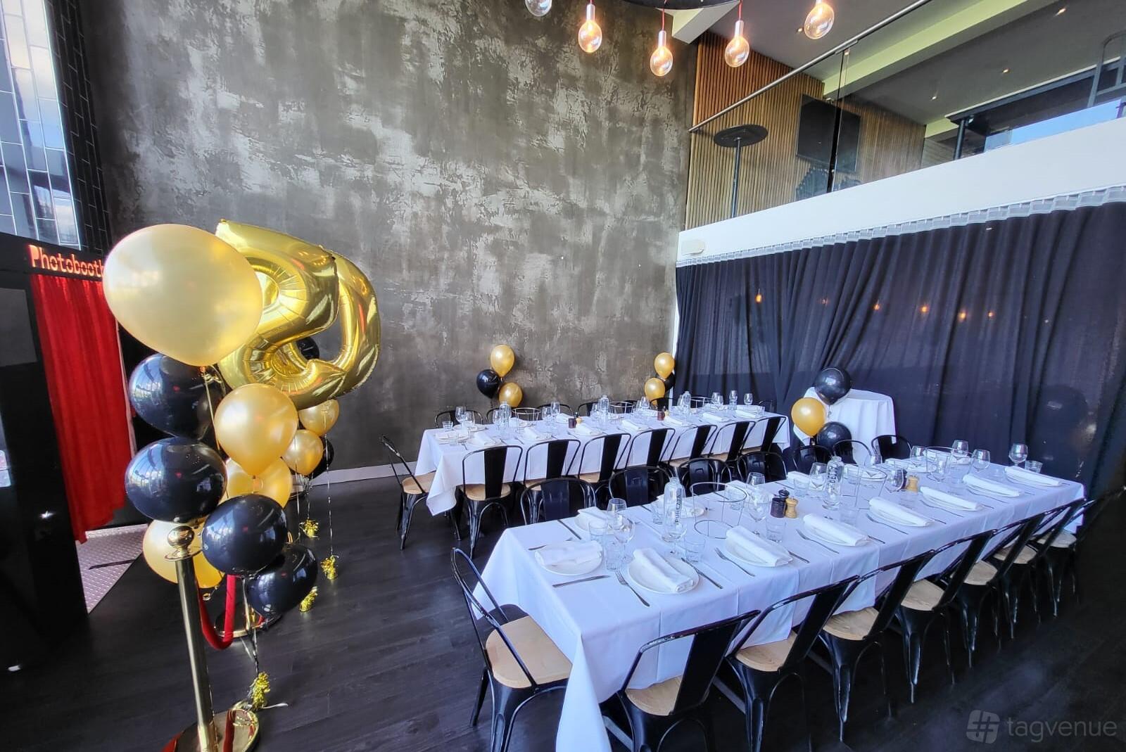 A cafe with long banquet tables, black and gold balloons, and floor-to-ceiling windows at The Last Piece Restaurant.