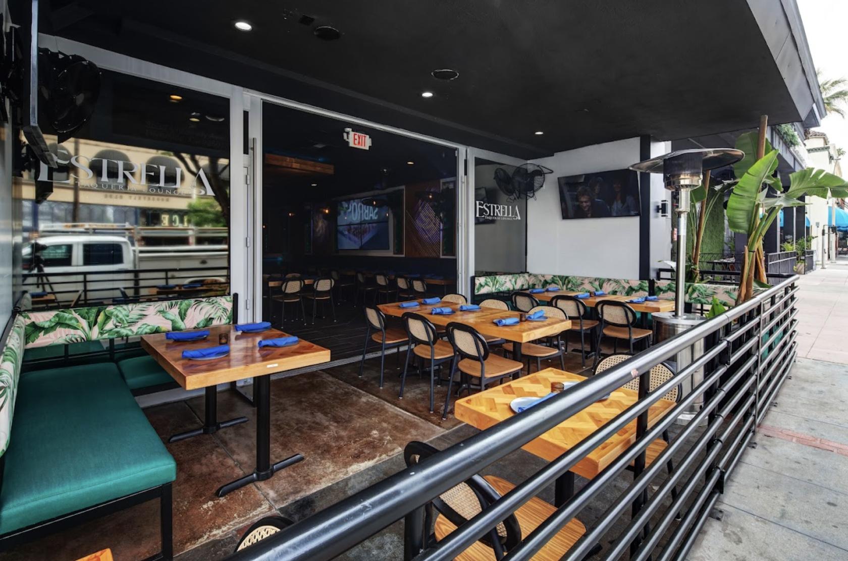 An outdoor restaurant patio with wooden tables, green cushioned bench seating, and potted plants at Estrella Taqueria Lounge.