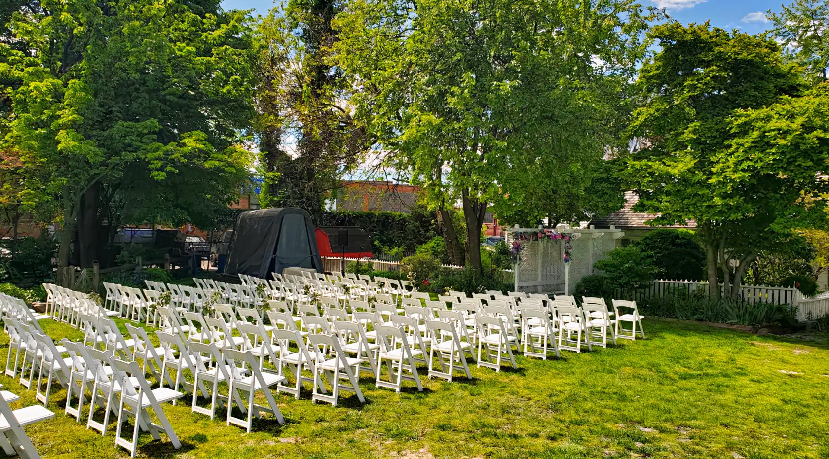 An outdoor event venue with rows of white folding chairs on a grassy lawn surrounded by trees at The Vander Ende - Onderdonk House.