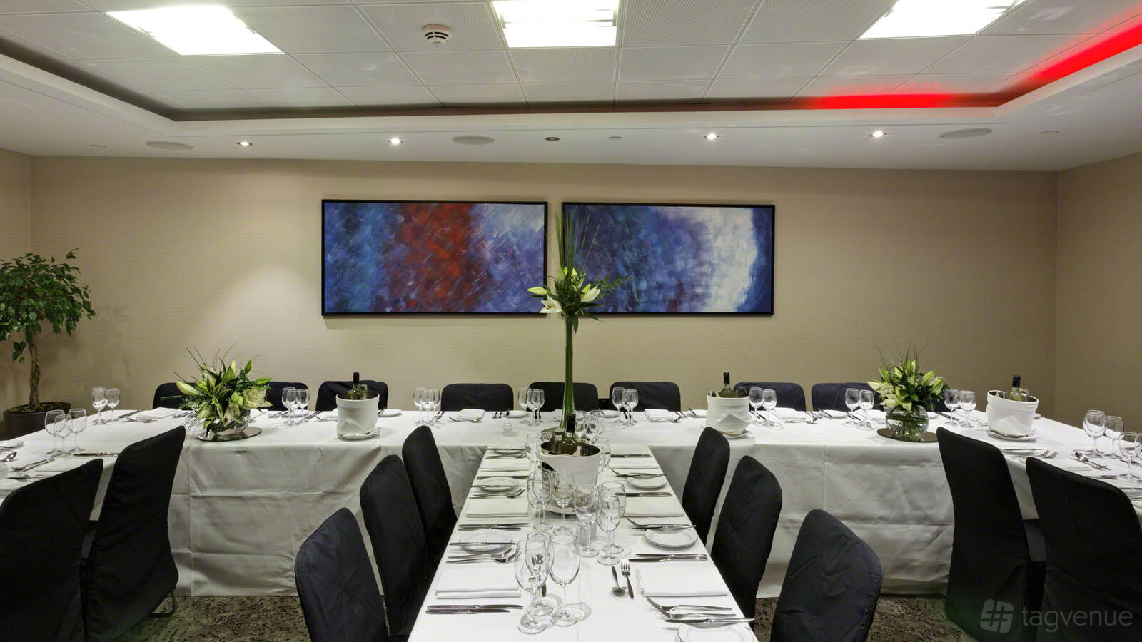 A function room with white-clothed banquet tables, black chair covers, and abstract wall art at Leonardo Royal London St Paul's.