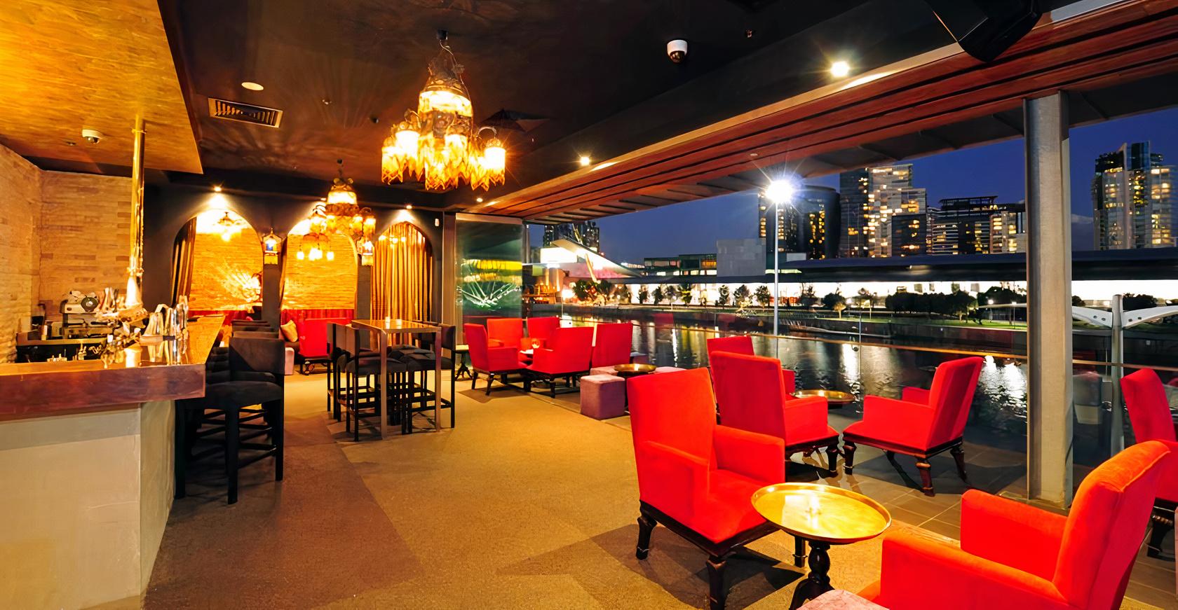 A terrace with red armchairs, high tables, decorative pendant lights, and river views at Byblos Bar and Restaurant.