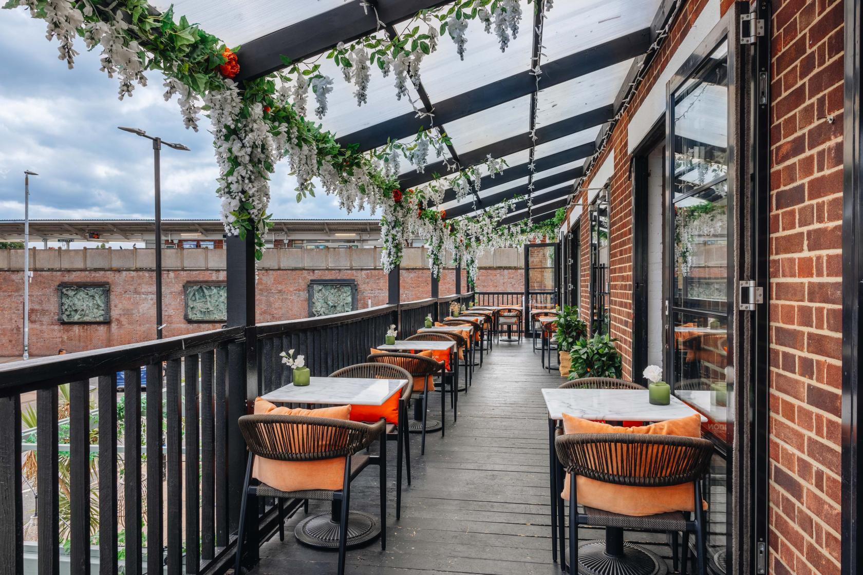 A covered balcony event space with hanging greenery, black railing, and small tables with orange chairs at Exhibit.