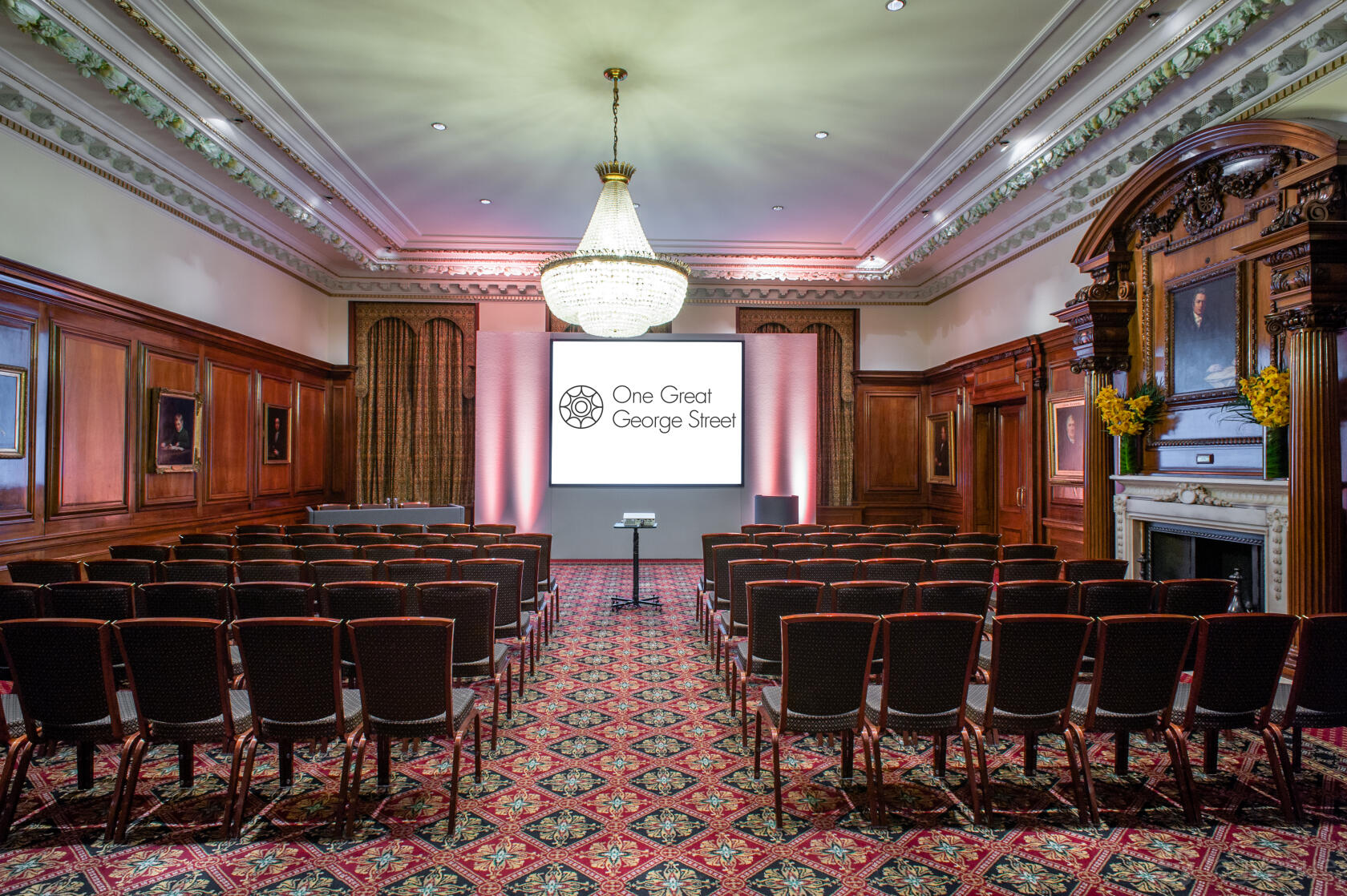 An event room with ornate wood paneling, a central chandelier, and theater-style seating at One Great George Street.