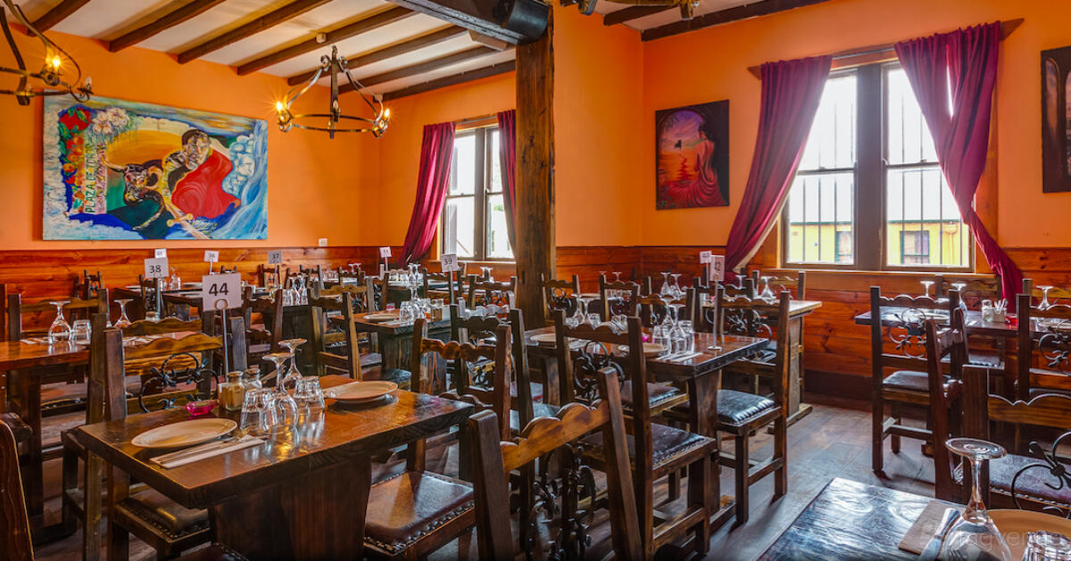 A restaurant dining room with wooden tables and chairs, colorful wall art, and large windows with red curtains at Spanish Tapas.