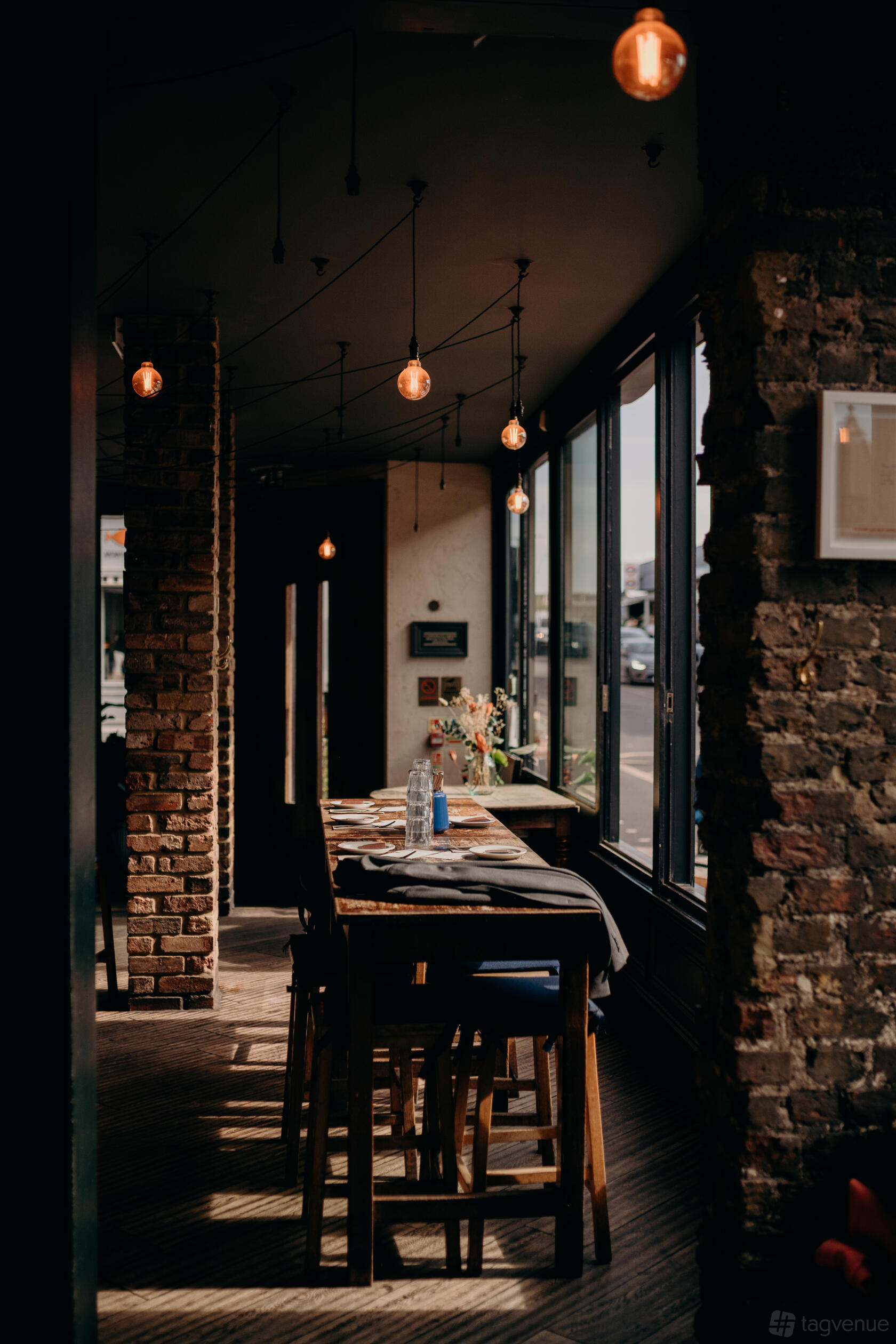 A bar with exposed brick walls, large windows, pendant lighting, and a wooden dining table at The Alice House Queen's Park.