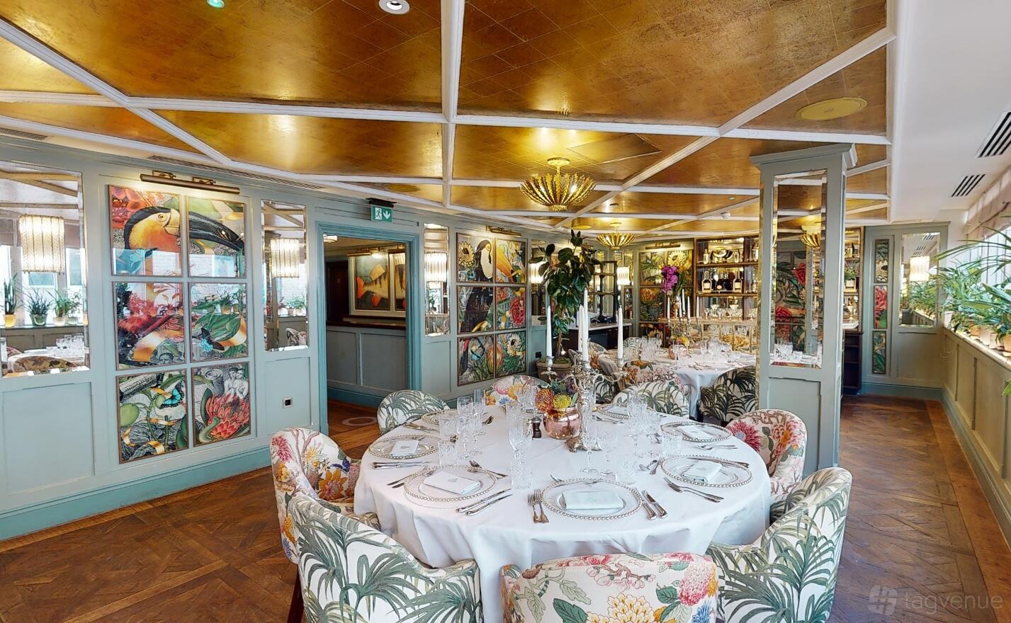 A dining room with round tables set with glassware, floral upholstered chairs, and botanical wall art at The Ivy Oxford Brasserie.