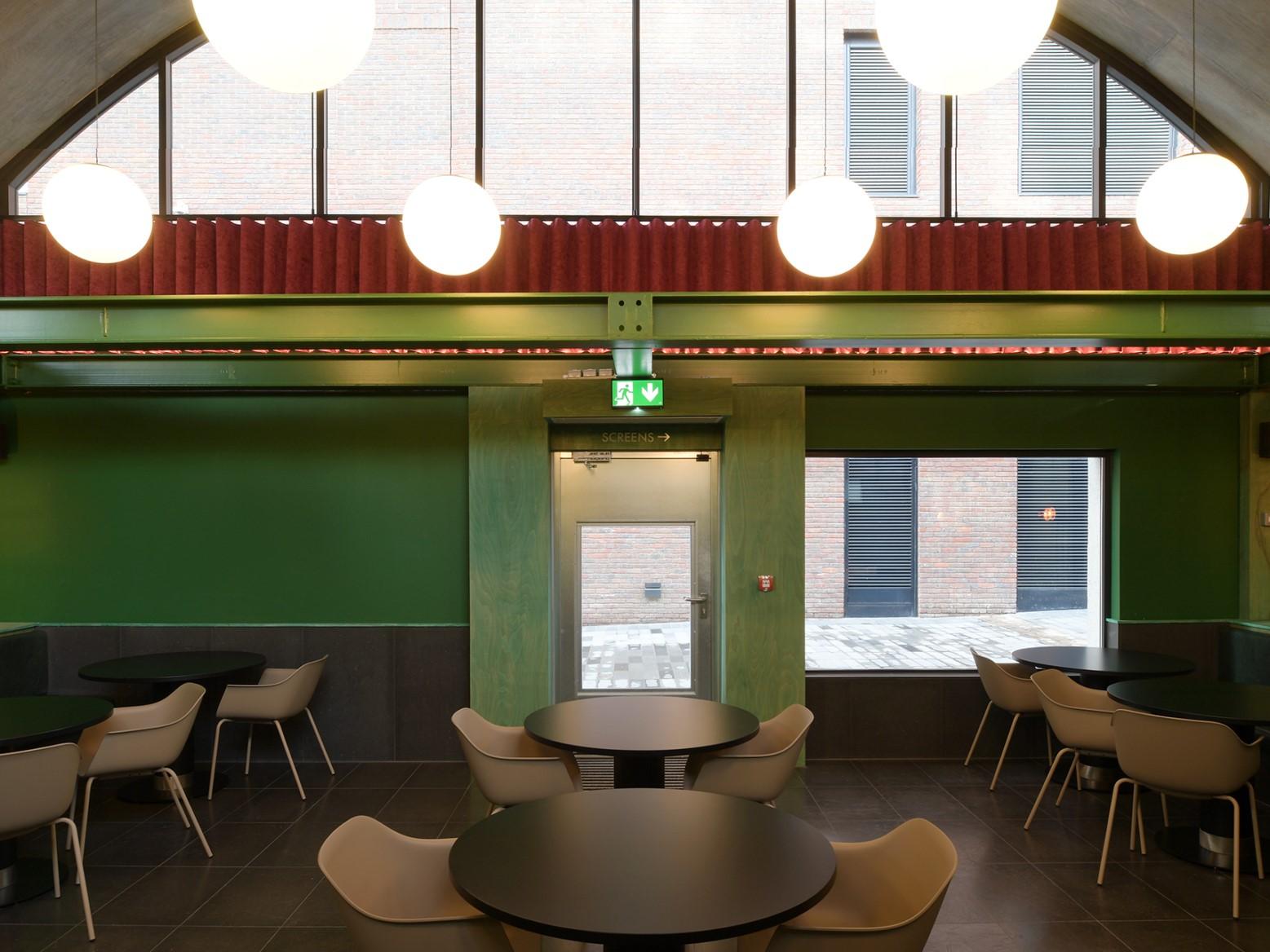 A cinema bar with round black tables, white chairs, globe pendant lights, and green walls at Curzon Camden.