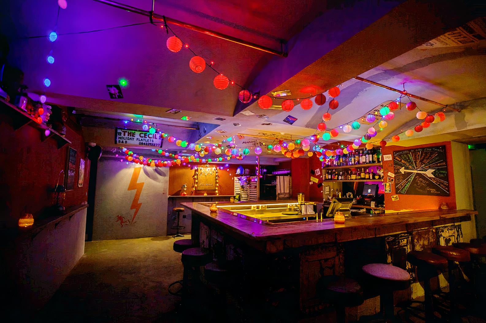An underground bar with colorful string lights, red lanterns, and a central counter at The Breakfast Club London Bridge.