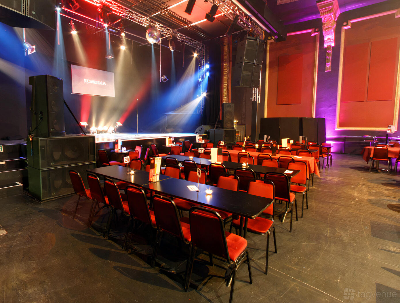 An auditorium with red chairs, rectangular tables, stage lighting, and a raised stage at Komedia.