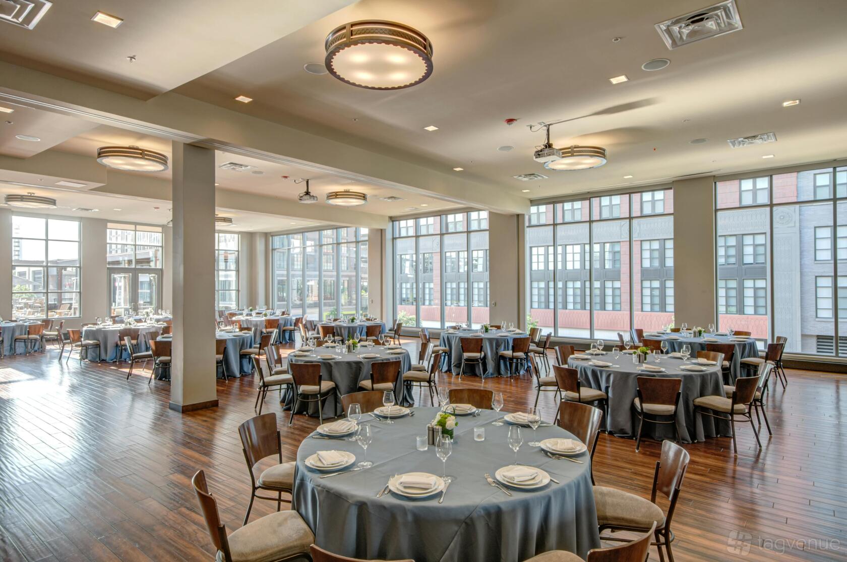 A ballroom with large windows, round tables with gray linens, and hardwood floors at Pinstripes - Bethesda.