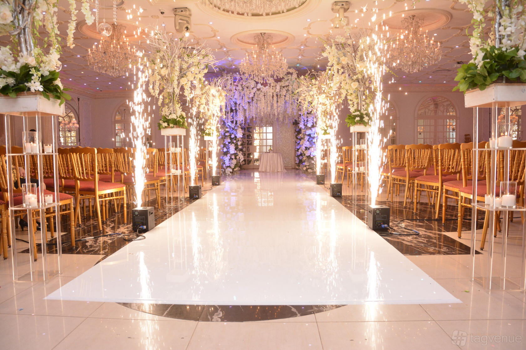 An event venue with a white aisle, gold chairs, floral arrangements, chandeliers, and indoor fireworks at Da Mikele Illagio.