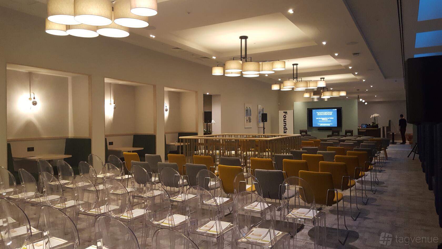 An event space with rows of transparent chairs, yellow armchairs, and ceiling lights at The Clubhouse 8 St. James's Square.