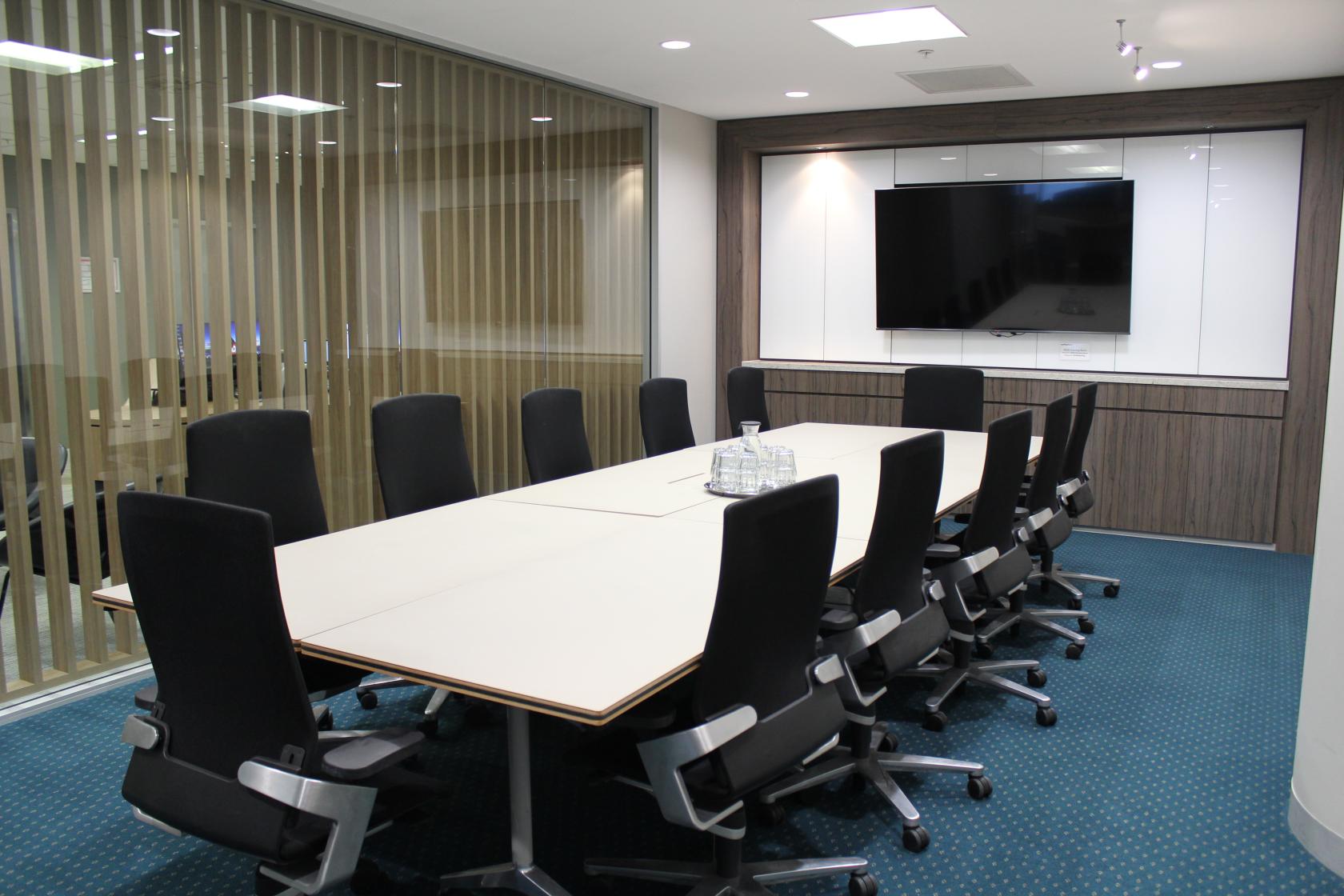 A meeting room with a long conference table, black swivel chairs, glass water set, and a wall-mounted screen at Workspace365 Parramatta.