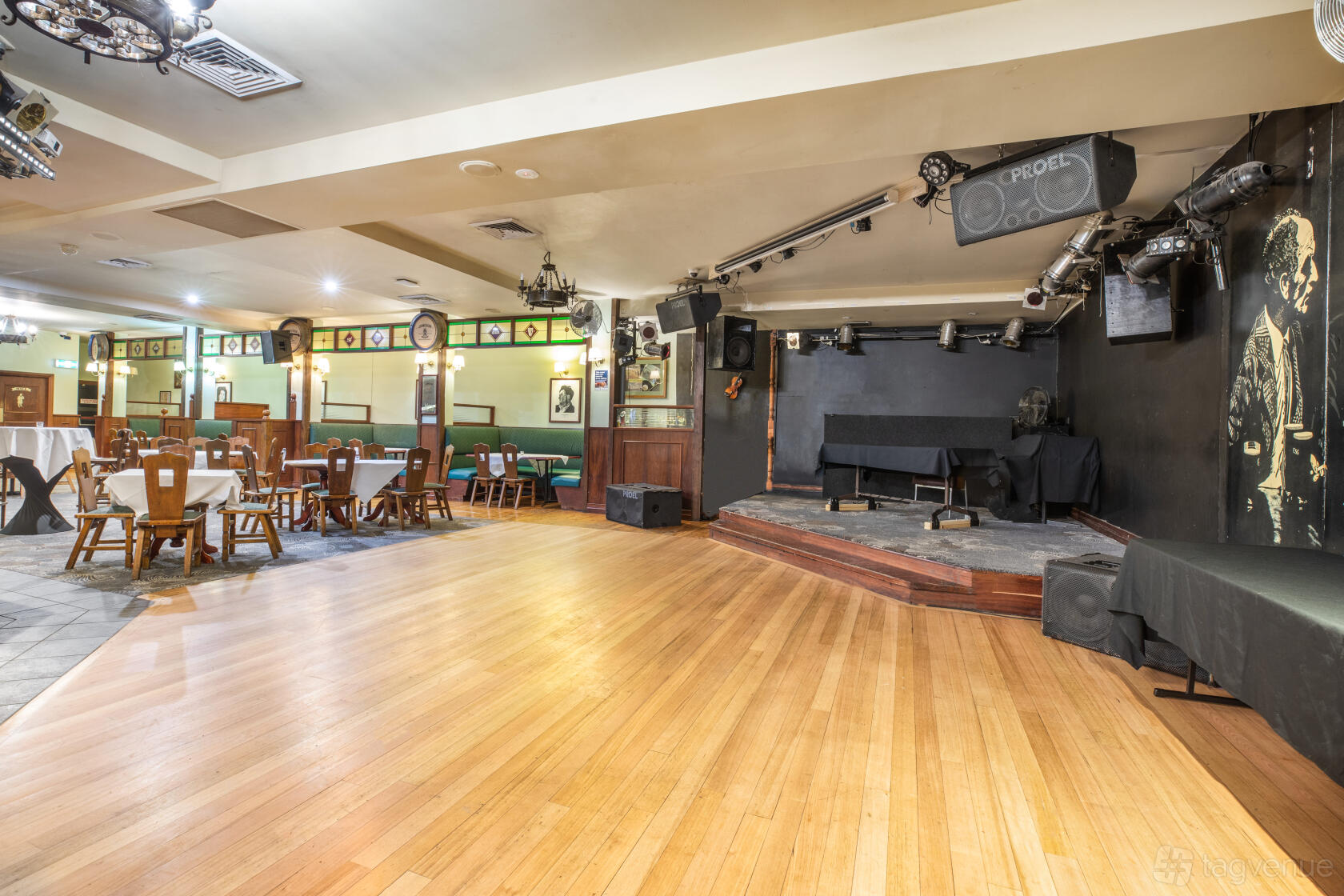 An event space in a pub with a wooden dance floor, stage with lighting, and round tables at Monash Hotel.
