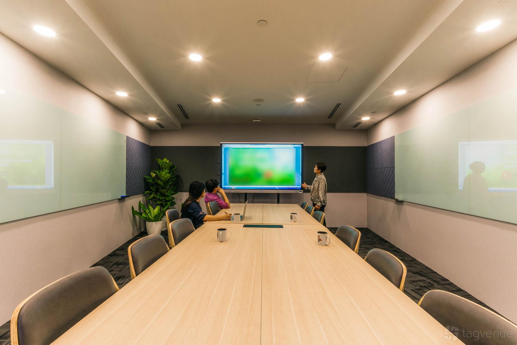 A meeting room with a long table, ergonomic chairs, dual glass whiteboards, and a large display screen at O2 Work @ Collyer Quay Centre.