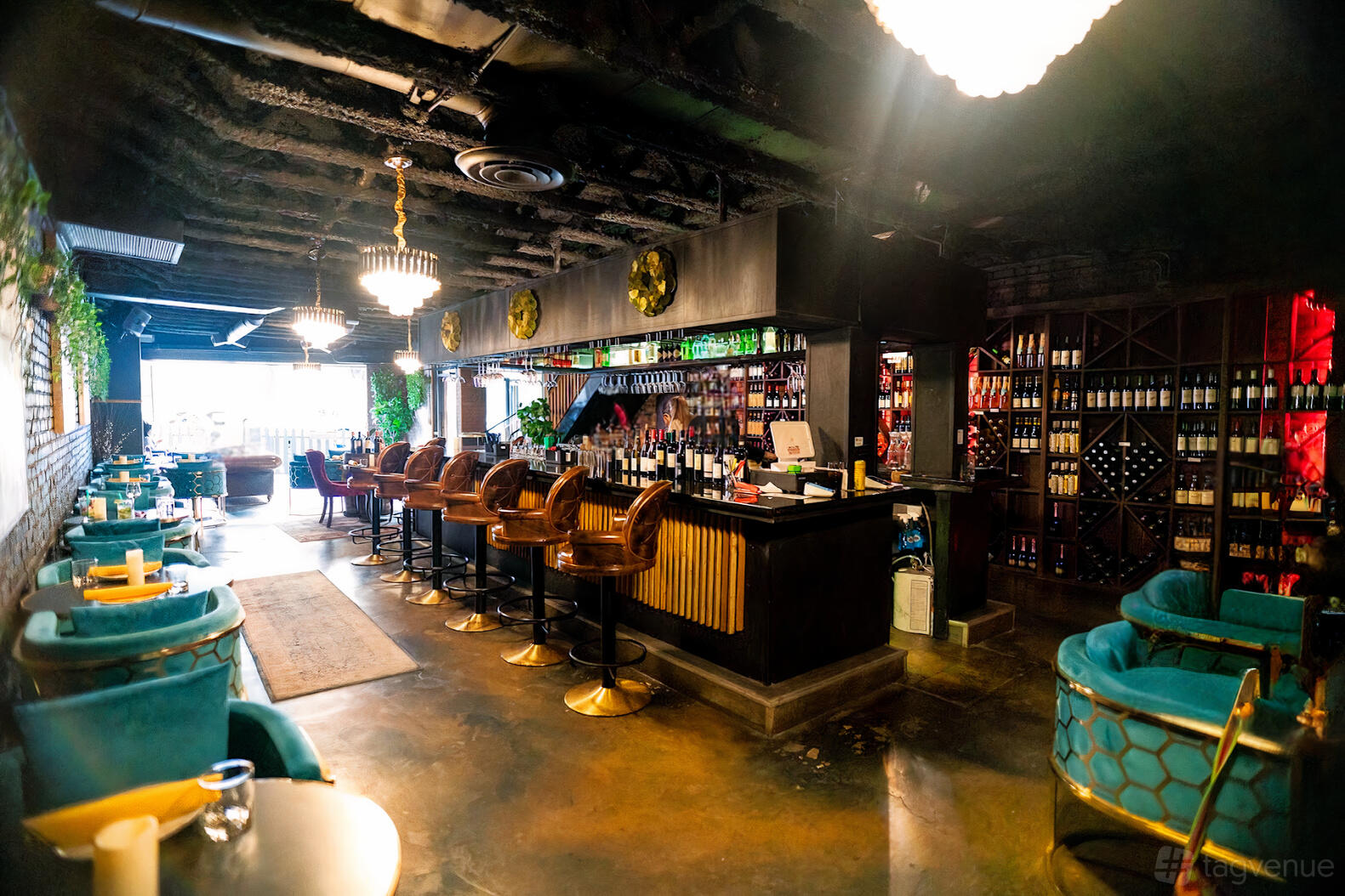 A bar with teal velvet chairs, a long counter, wine bottle displays, and chandeliers at Barrel Cheese & Wine.