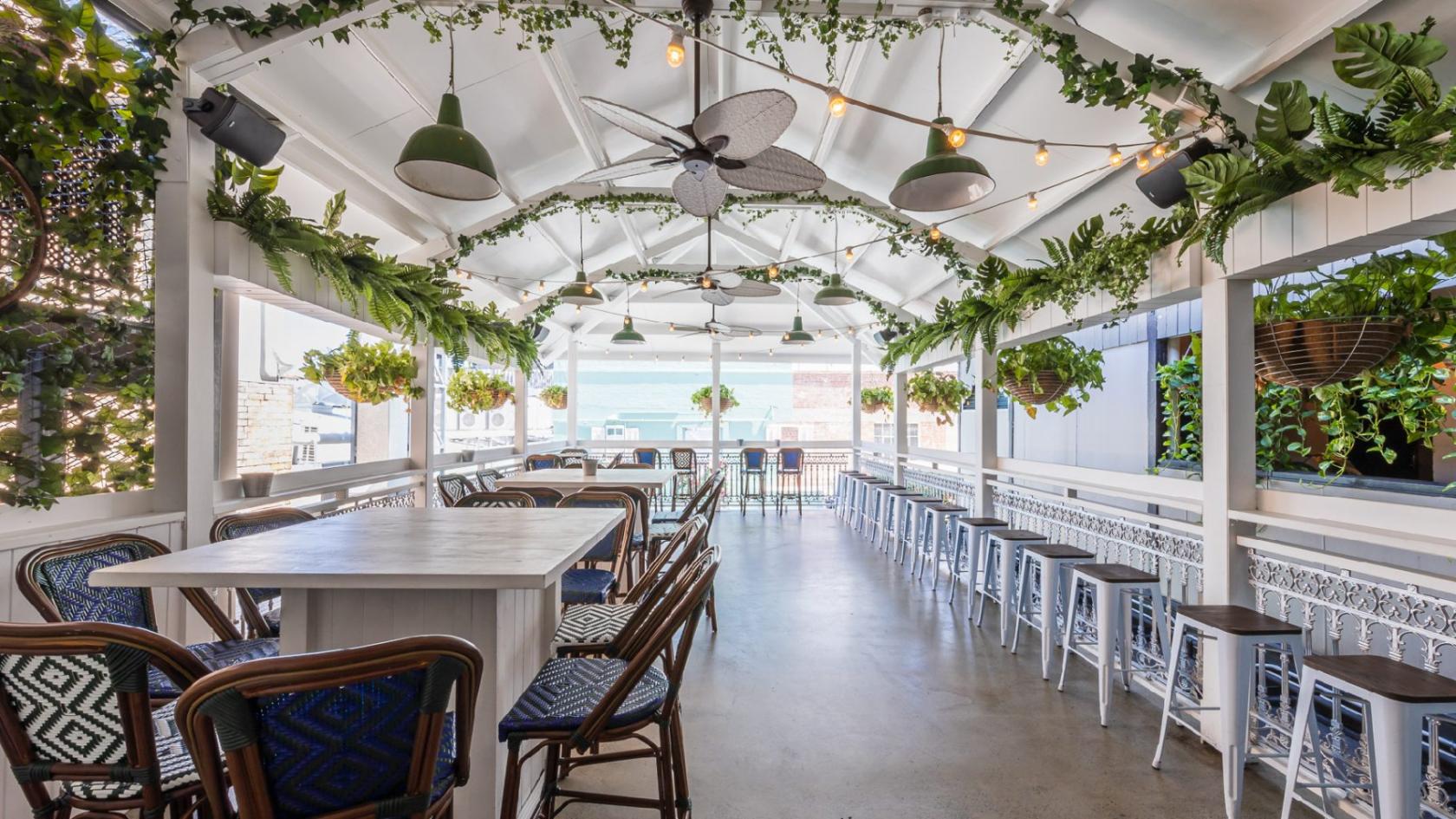 A covered terrace pub with hanging plants, ceiling fans, white bar stools, and leafy decor at The Prince Consort.
