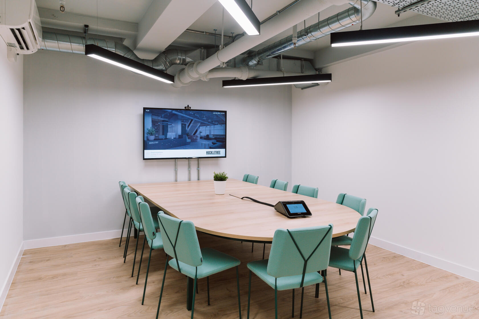 A meeting room with a large oval table, mint green chairs, a wall-mounted screen, and exposed ductwork at Huckletree Soho.