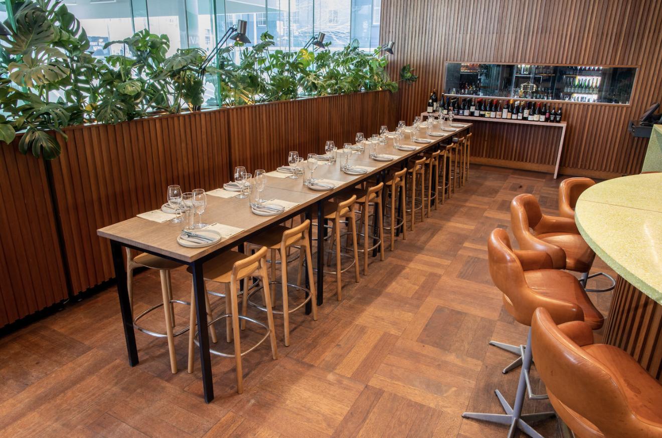 An event space in a bar with long wooden tables, barstools, potted plants, and a selection of wines at Tiny's.