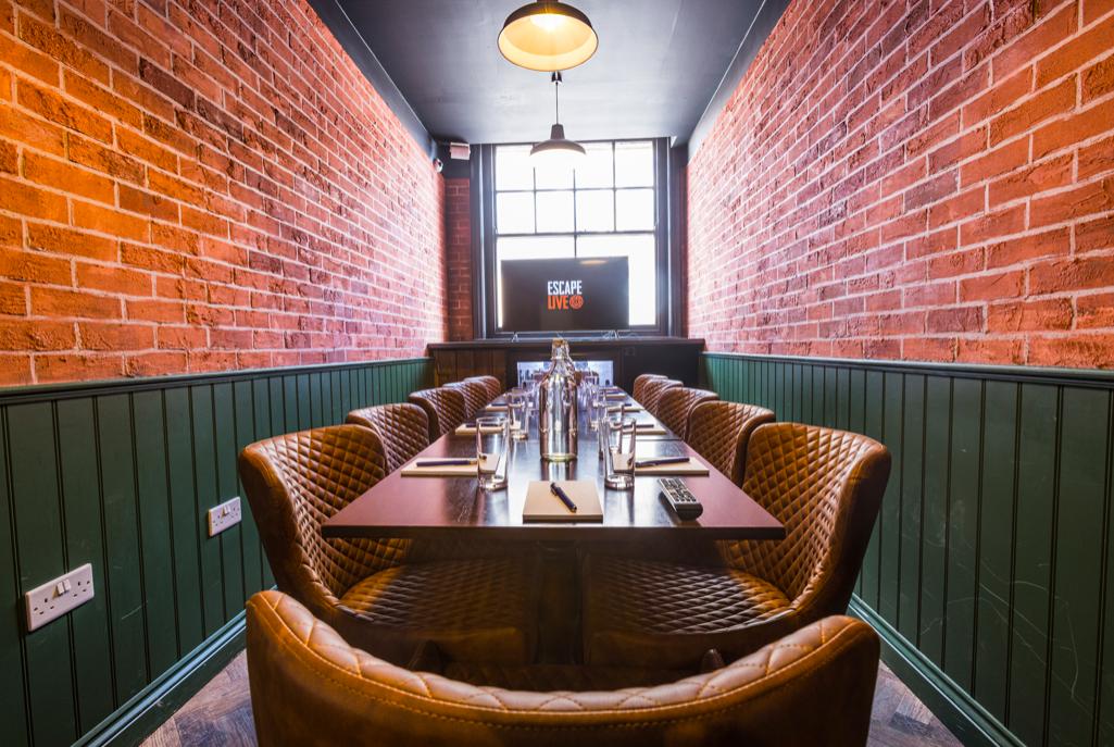A bar with exposed brick walls, leather chairs, and a long dining table set under pendant lights at Escape Live Birmingham.