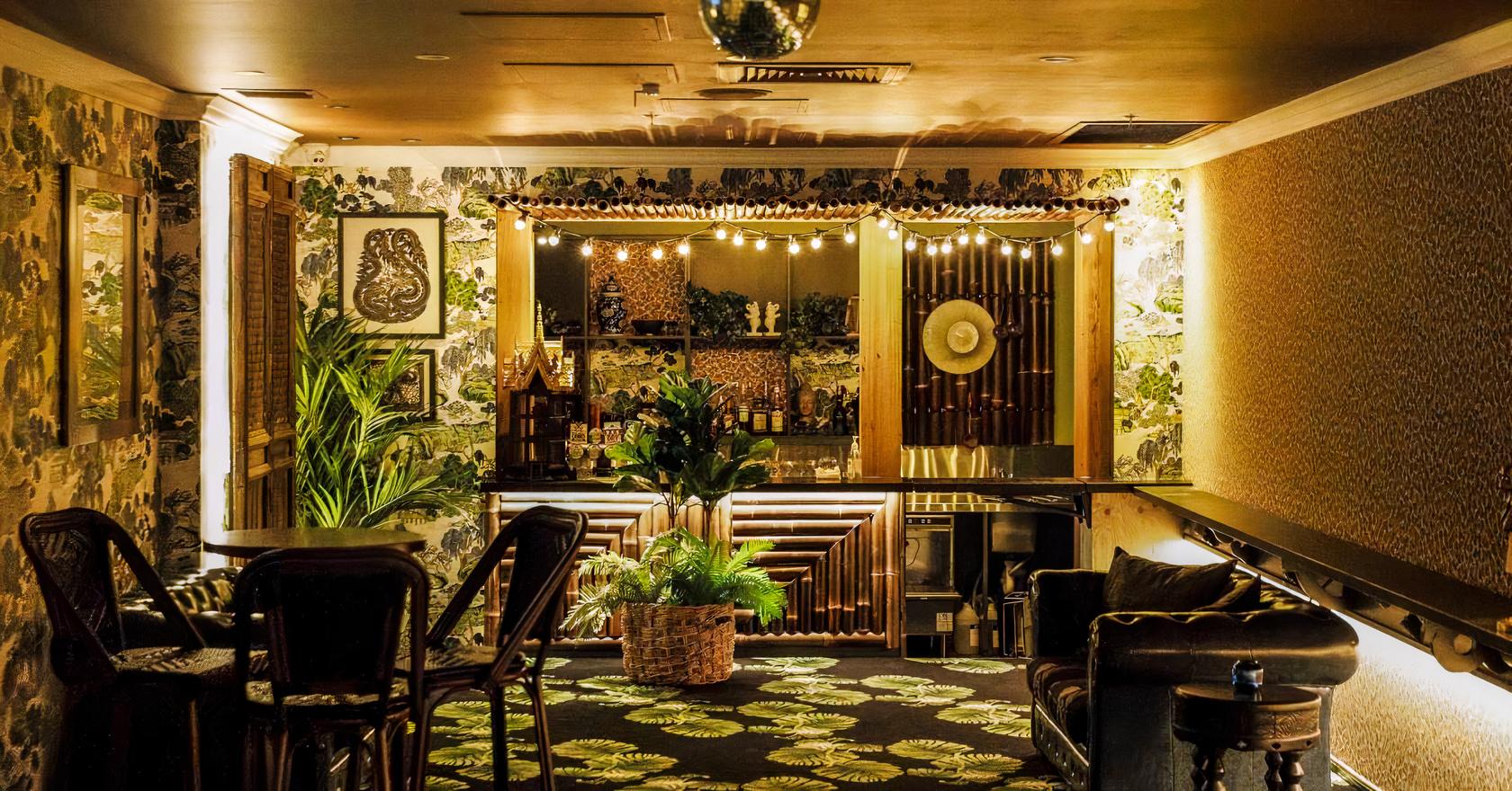 A function room in a pub with a patterned carpet, botanical wallpaper, string lights over a wooden bar, and leafy plants at The Prince Consort.
