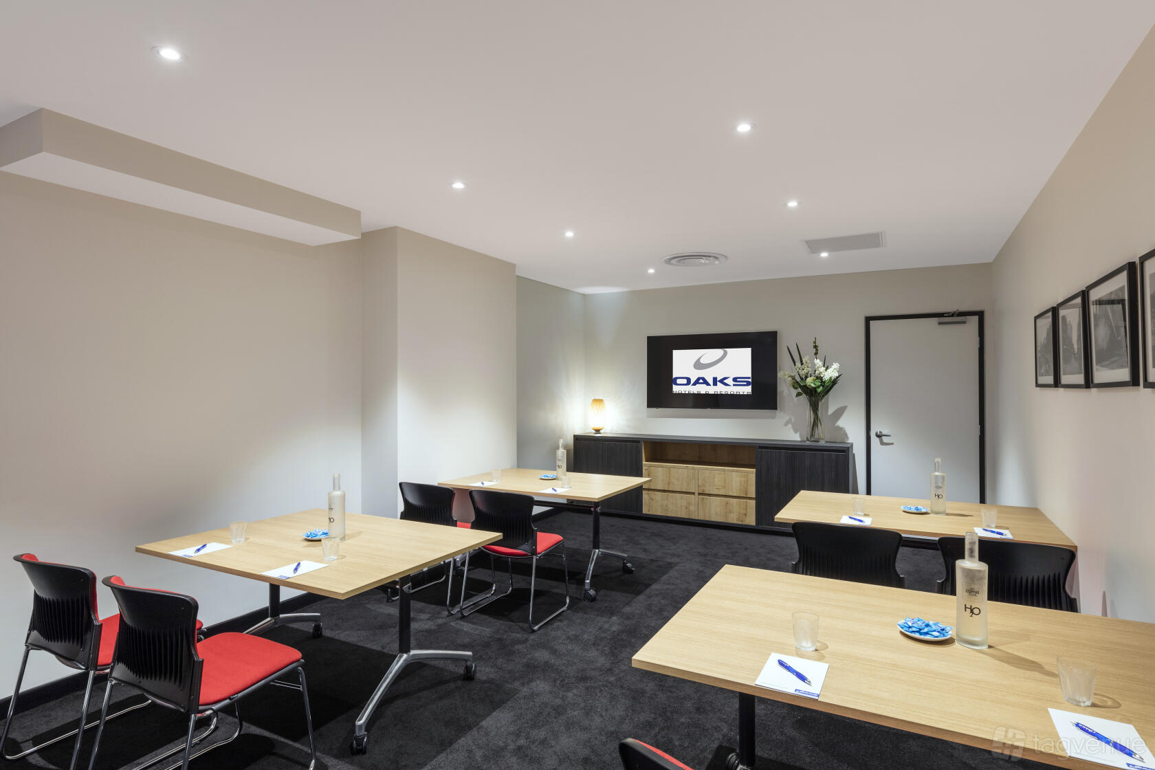 A meeting room with rectangular tables, black chairs, a wall-mounted screen, and minimalist decor at Oaks Embassy.