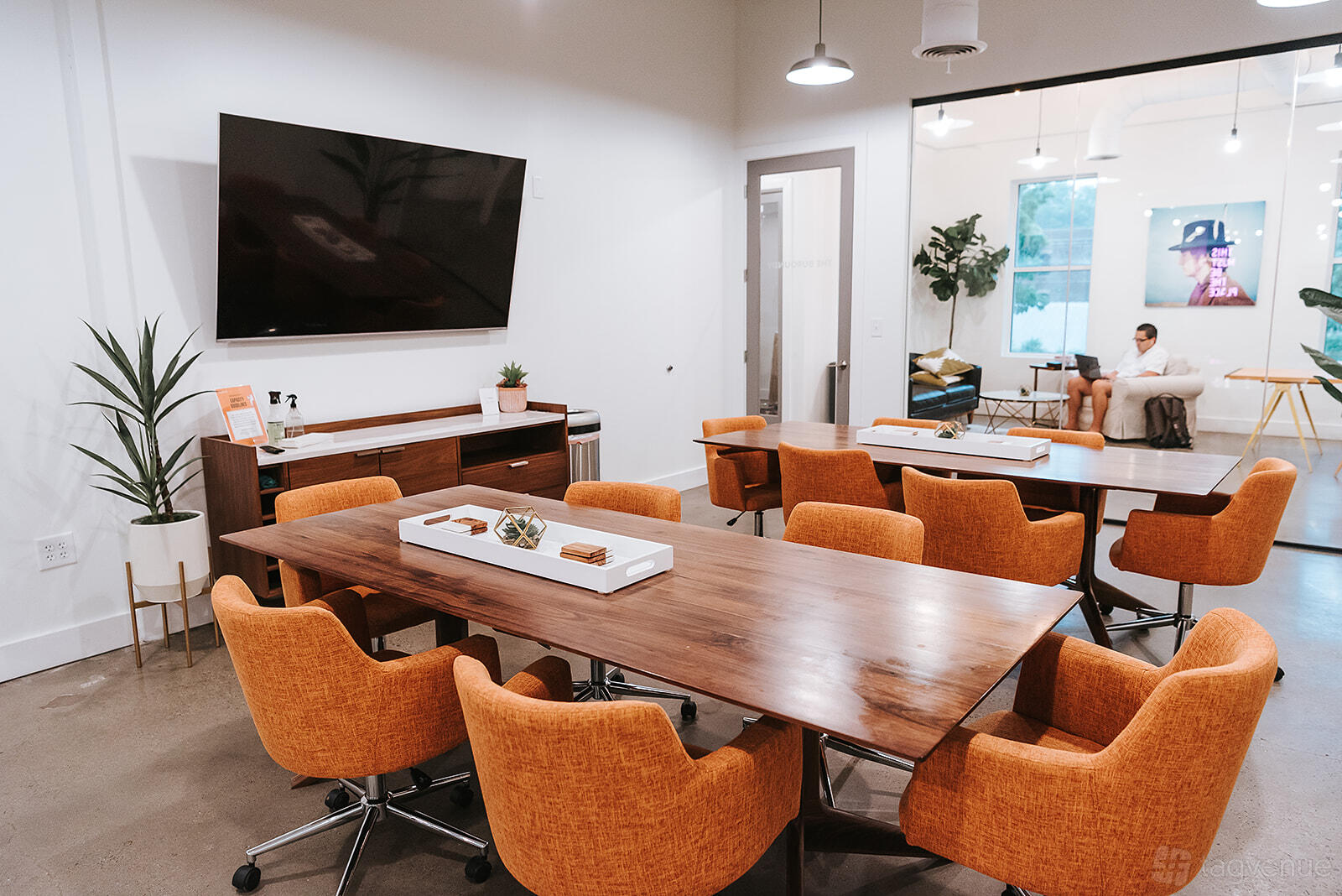 A coworking space with wood tables, orange chairs, a wall-mounted TV, and potted plants at THRIVE - Alpharetta.