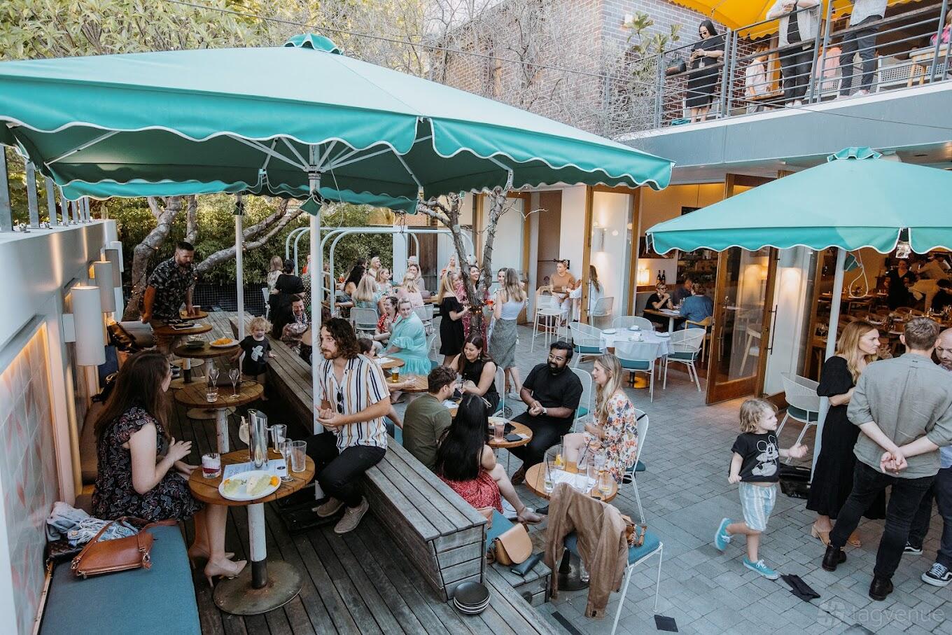 A terrace at a pub with large green umbrellas, wooden benches, and people dining at The Tilbury Hotel.