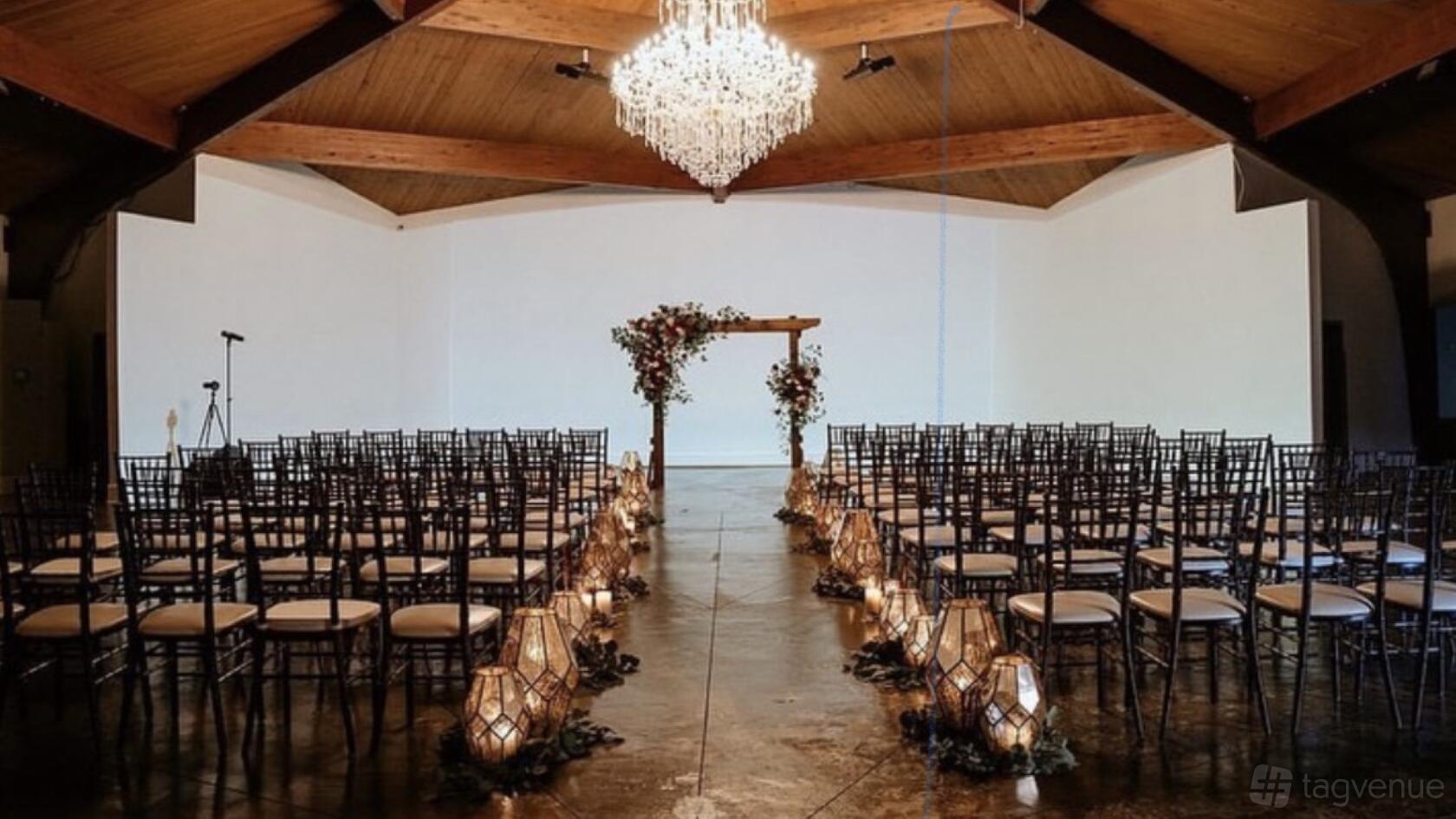 A hall with a central aisle lined by lanterns, dark wooden chairs, and a crystal chandelier at The West Venue.