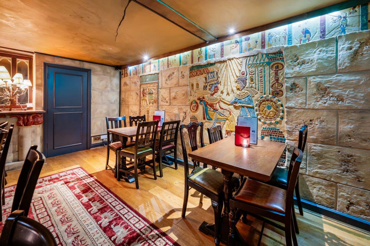 A pub dining room with Egyptian-style wall murals, wooden tables, and a patterned rug at Bacchus.