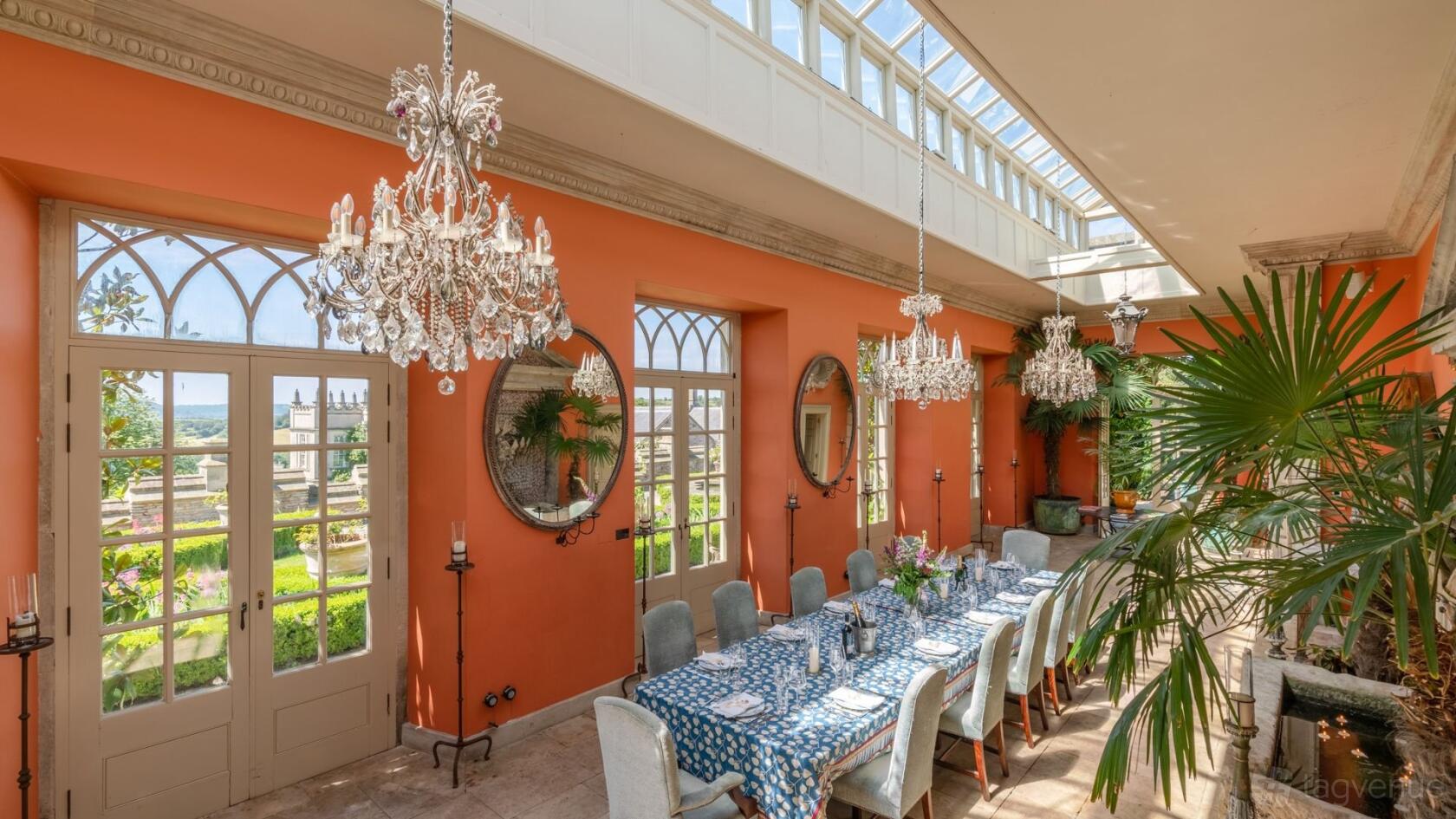 An event venue with crystal chandeliers, large arched windows, and a long dining table in THE ORANGERY at Euridge Weddings.